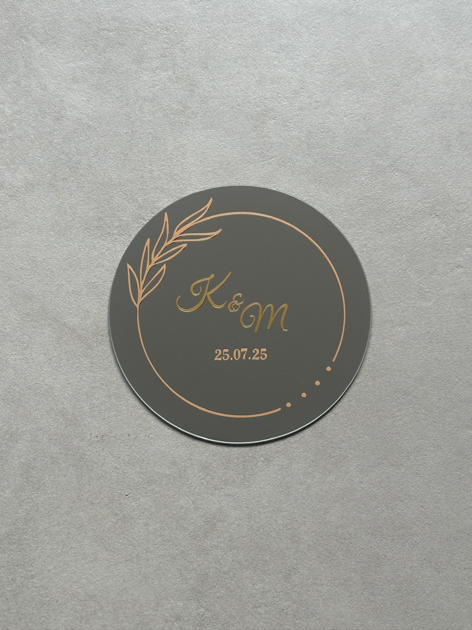 Spiegel met sierlijke voorletters en datum in het midden. Daar omheen een dunne cirkel die met blaadjes en drie stipjes wordt onderbroken.